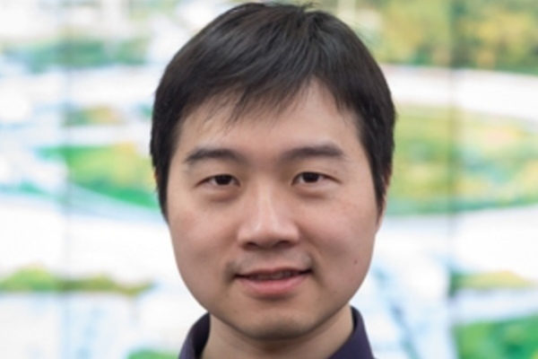 A man with dark hair and a gentle smile, wearing a dark collared shirt. Blurred green background.